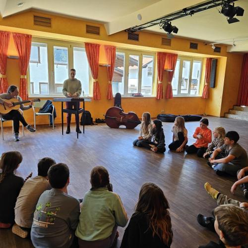 Kinder sitzen im Kreis am Boden und zwei Frauen spielen auf einer Gitarre und einem Hackbrett