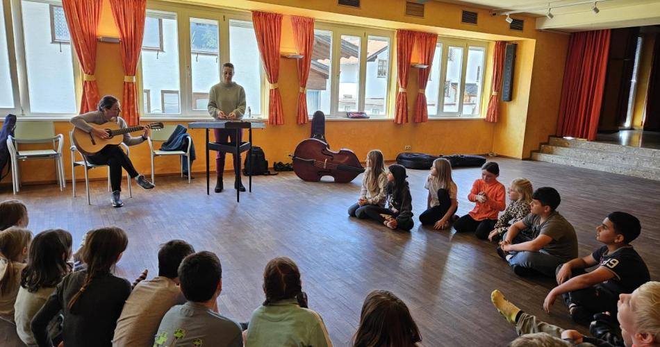Kinder sitzen im Kreis am Boden und zwei Frauen spielen auf einer Gitarre und einem Hackbrett