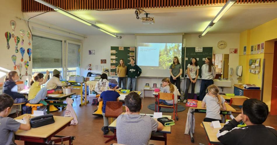 5 Mädchen stehen vorne an der Tafel und die Kinder blicken nach vorne zu den Mädchen